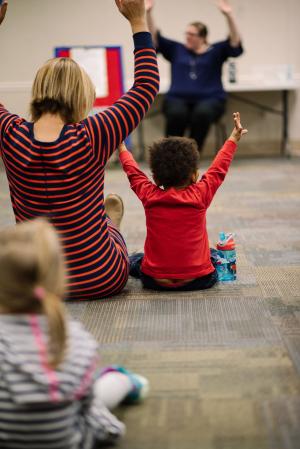 A child and mother mimic a librarian's arm motions during a storytime game